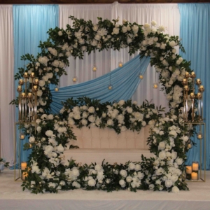 A stunning stage setup featuring a light blue drape backdrop, a floral arch adorned with white roses and greenery, and elegant gold candelabras