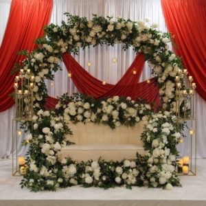 A beautiful floral arch adorned with white roses and greenery, set against a red drape backdrop on stage, with matching gold candelabras adding elegance.